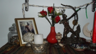 This is the shelf below the ancestor shrine, and we have Hermes and Artemis here. The jar with the coins and the small silver dish in front of His picture belong to Hermes. The white candle is for Artemis, representing the full moon. The serpent and the dish in front of Artemis' statue are Artemis' things. The dish contains the prayer beads I made for Her. Hanging at the back is a cingulum I made some time ago.
