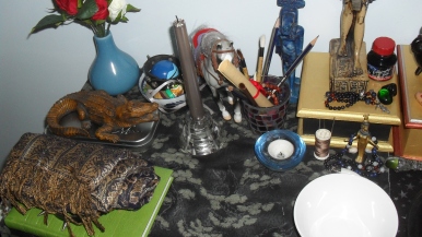 Close-up of Sobek's side of the shrine. The green book is my ritual/prayer book, and on top of that is my collar and prayer shawl. Behind that is my calligraphy box, which Sobek is protecting. The small bowl between the horse and the blue vase contains trinkets and other assorted things. The blue ball in the cuff is a Dive Ball with a Totodile in it. There are more calligraphy things in the candle holder next to the horse. Have a better look at Nit's shrine in front of Sobek's main statue.